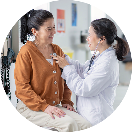 An Asian doctor holds a stethoscope up to the chest of her patient, a white woman with a black ponytail and orange sweater.