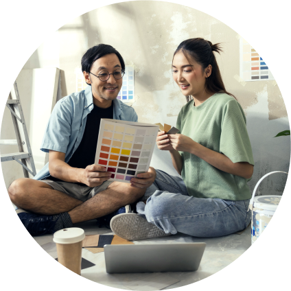 While painting their new home, a young Asian couple sits cross-legged on the floor smiling and looking at color swatches.