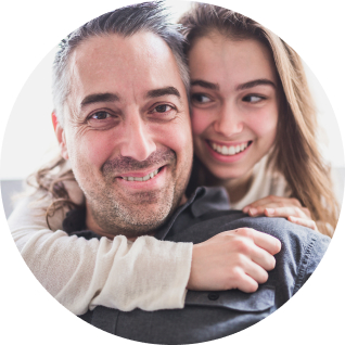 Man smiles at camera while woman behind him smiles at him with her arm around his neck