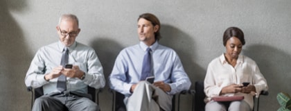 Mature man, young man, and woman in business attire reading smartphones while sitting in a waiting area