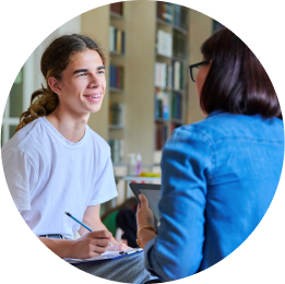 A smiling teenager with long hair in a ponytail engages with their therapist, a woman in glasses whose back is to us.