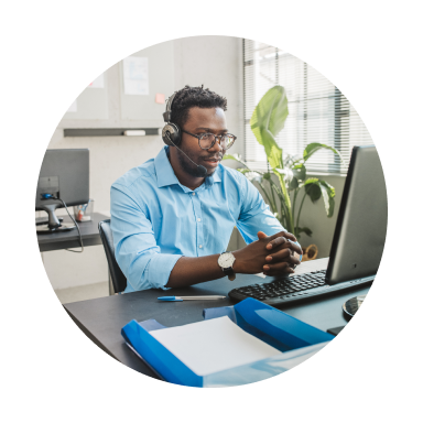 A young Black customer service representative wears glasses, a blue shirt and a headset while looking at a computer screen.