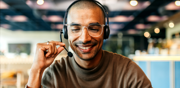 A young man, wearing glasses and a headset, laughs as he looks down toward his desk.