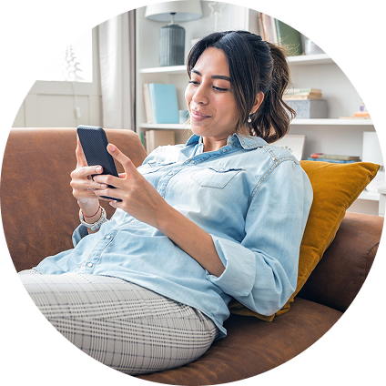 A woman with straight dark hair smiles as she leans back on her couch and looks at her phone.