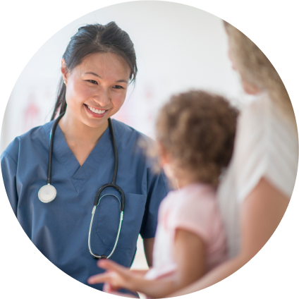 A young Asian doctor in blue scrubs smiles at her patient, a young girl sitting on her mother's lap.